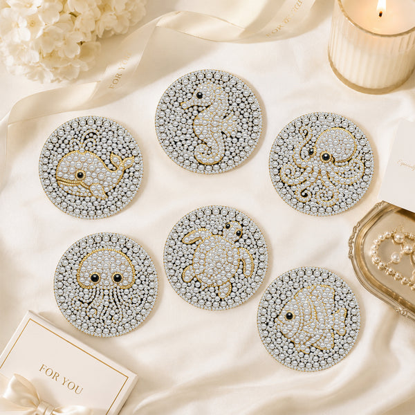 Six round coasters with sea creature designs, including a whale, octopus, crab, fish, and seahorse, decorated with white rhinestones and gold outlines, arranged on a cream satin fabric with decorative props.