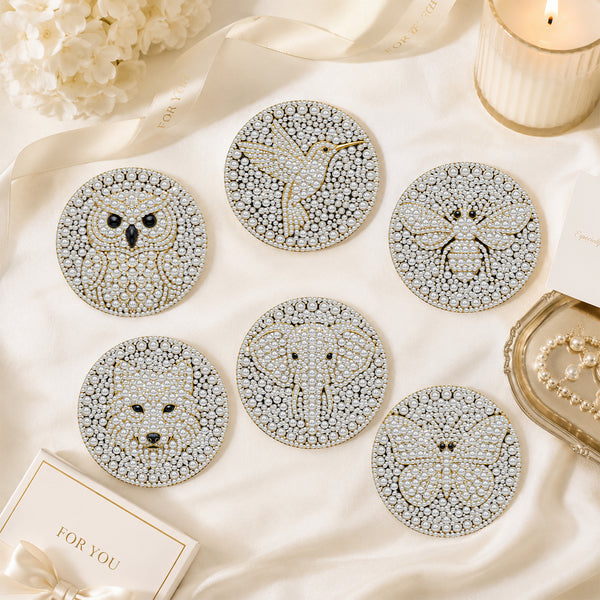 Six round beaded coasters featuring animal and nature designs including an owl, hummingbird, bee, fox, elephant, and butterfly, arranged on a cream satin fabric with decorative props.