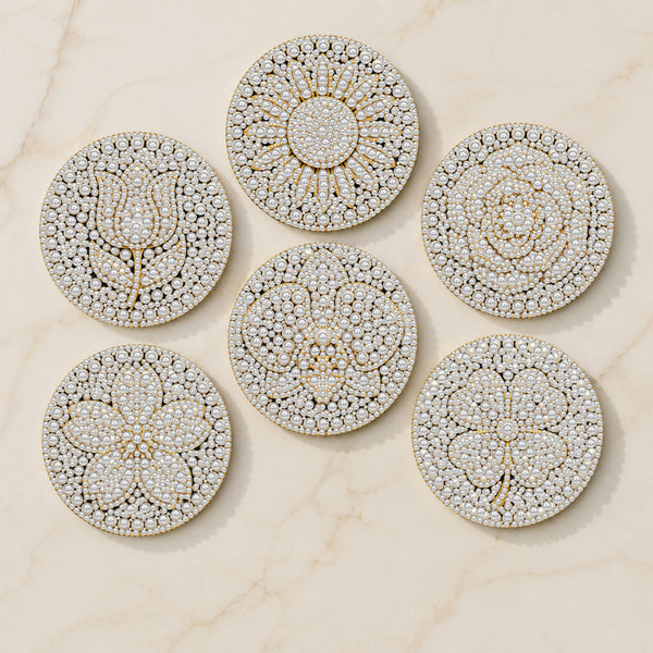 Six round gold-toned brooches embellished with white stones in floral and geometric patterns, arranged on a light beige marble surface.