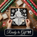 A square gift box wrapped in black wrapping paper with a white snowman and snowflake pattern, tied with a wide silver ribbon and bow. Two hands are holding the front of the package.