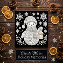 A square black-and-white holiday art print featuring a knitted snowman holding a lantern, surrounded by snowflakes and branches. The print is displayed on a dark wood surface with pinecones, dried orange slices, fairy lights, and a banner reading 'Create Warm Holiday Memories'.