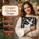 A woman in a cream knit sweater holds a black square Christmas ornament or plaque with a white reindeer and snowflake design. The background is a warm, blurred holiday scene with lights and pine branches.