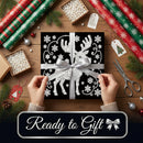 A square black gift box wrapped in a silver ribbon with a white reindeer and snowflake pattern, held by two hands over a wooden table decorated with wrapping paper, scissors, and pine branches. A dark banner at the bottom reads 'Ready to Gift' with a silver bow icon.