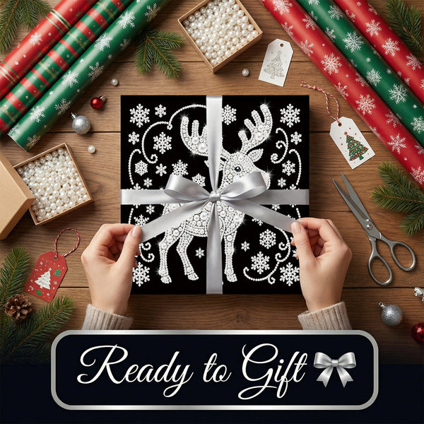A square black gift box wrapped in a silver ribbon with a white reindeer and snowflake pattern, held by two hands over a wooden table decorated with wrapping paper, scissors, and pine branches. A dark banner at the bottom reads 'Ready to Gift' with a silver bow icon.