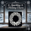 A square black canvas or box featuring a white glittery snowflake and wreath design, displayed in front of snowy window panes with the text 'Sparkling Holiday Charm' above it.