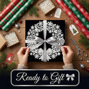 A wrapped gift box with a black background and white floral snowflake pattern, tied with a silver ribbon, held by two hands over a wooden table decorated with wrapping paper, pine branches, ornaments, and scissors. A banner at the bottom reads 'Ready to Gift'.