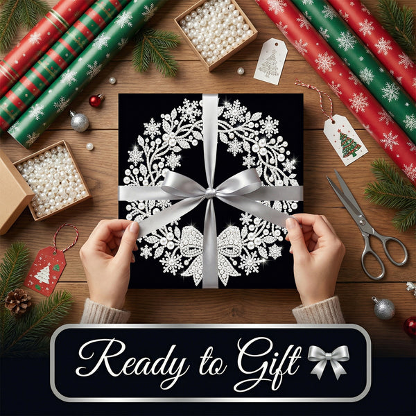 A wrapped gift box with a black background and white floral snowflake pattern, tied with a silver ribbon, held by two hands over a wooden table decorated with wrapping paper, pine branches, ornaments, and scissors. A banner at the bottom reads 'Ready to Gift'.