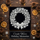 A square black print or canvas featuring a white glittery wreath with snowflakes and a bow, styled on a dark wood surface with dried orange slices, pinecones, fairy lights, and silver snowflake decorations. Text below reads 'Create Warm Holiday Memories'.