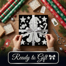 A square gift box wrapped in black wrapping paper with white holiday illustrations, tied with a wide silver ribbon and bow, held by two hands over a wooden tabletop. The scene is decorated with rolls of red and green wrapping paper, pine branches, gift tags, scissors, and small ornaments, with a banner at the bottom reading 'Ready to Gift'.
