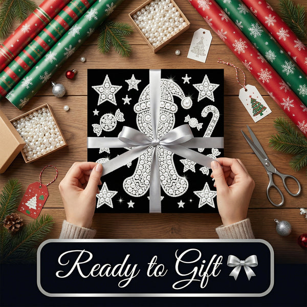A square gift box wrapped in black wrapping paper with white holiday illustrations, tied with a wide silver ribbon and bow, held by two hands over a wooden tabletop. The scene is decorated with rolls of red and green wrapping paper, pine branches, gift tags, scissors, and small ornaments, with a banner at the bottom reading 'Ready to Gift'.