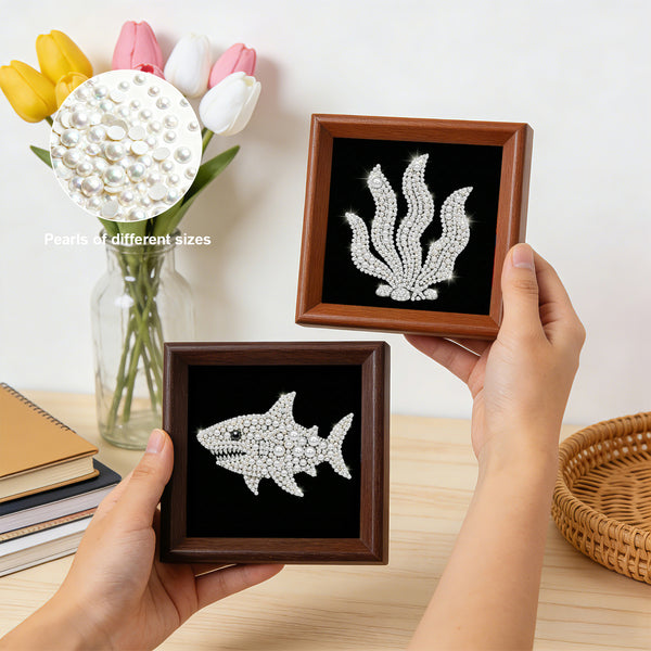 Two decorative brooches in wooden frames held by hands on a table with flowers and books.