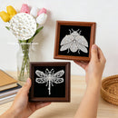 Two hands hold small framed diamond painting kits showing completed beetle and dragonfly designs on black backgrounds. A circular inset highlights white pearls of different sizes.