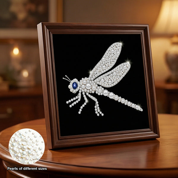 A framed dragonfly embellishment made of white pearls and clear rhinestones, with a blue gemstone eye, displayed on a black background inside a dark wood frame. The frame sits on a wooden table in a warmly lit room, and a circular inset shows the texture of the pearls.