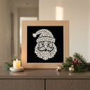 A framed Christmas diamond painting or mosaic art piece depicting Santa Claus's face and hat on a black background, set in a light wood frame and displayed on a wooden cabinet with holiday decorations including a candle, pine branches, and ornaments.