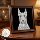A framed portrait artwork of a dog made from white pearls and silver rhinestones on a black background, displayed on a wooden table in a warmly lit room. A circular inset shows pearls of different sizes.