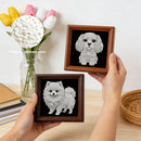 Two hands hold small framed diamond painting or rhinestone pet art pieces showing white dog designs on black backgrounds, with wooden frames. A vase of tulips, books, and a woven tray sit on a table behind, and a circular inset highlights pearl texture.