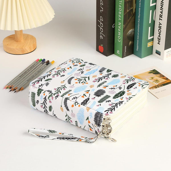 A hardcover notebook with a white cover decorated with a colorful botanical leaf pattern, shown on a white desk next to a pleated lamp, colored pencils, and books.