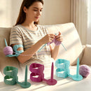 A woman sits on a beige sofa knitting with light purple yarn and metal needles, supported by a light blue arm knitting frame. Four additional knitting frames in green, magenta, and light blue are arranged on a table in front of her.