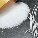 White plastic pellets spilled from a gold-colored bag onto a dark gray textured surface, with several white rods or sticks placed on the right side.
