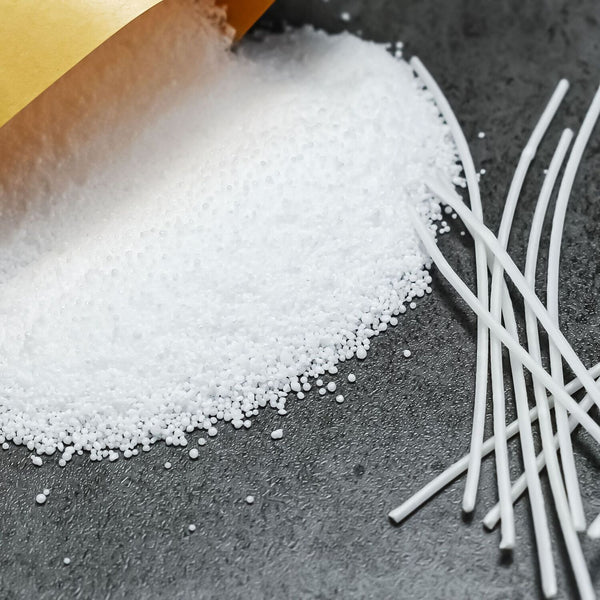 White plastic pellets spilled from a gold-colored bag onto a dark gray textured surface, with several white rods or sticks placed on the right side.