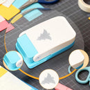 A small white and light blue craft punch tool with a gray shark logo, shown on a dark gridded work surface with scissors and colorful paper pieces around it.