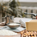 A styled kitchen or craft workspace scene with a stainless steel bowl filled with white rice on a digital scale, a metal pitcher, a cardboard box, and a pile of brown nuts on a wooden surface, with a blurred indoor background.
