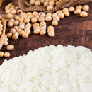 White soy wax flakes in the foreground with scattered soybeans and a wooden satchel in the background on a dark wood surface.