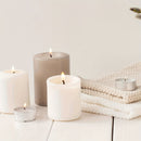 A set of lit pillar candles and tealight candles in neutral tones, including white, warm off-white, and taupe, arranged on a white wooden surface next to a folded cream knit blanket.