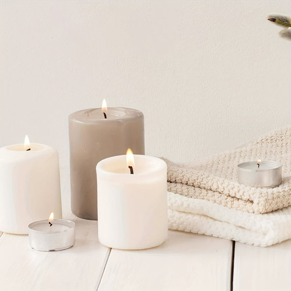 A set of lit pillar candles and tealight candles in neutral tones, including white, warm off-white, and taupe, arranged on a white wooden surface next to a folded cream knit blanket.