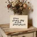 Decorative sign with 'live every day with intention' text on a wooden surface with dried flowers.