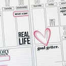 A close-up of a weekly planner page with lined sections, black printed text, and pink handwritten accents, including a heart outline and the words 'REAL LIFE' and 'goal getter.'.