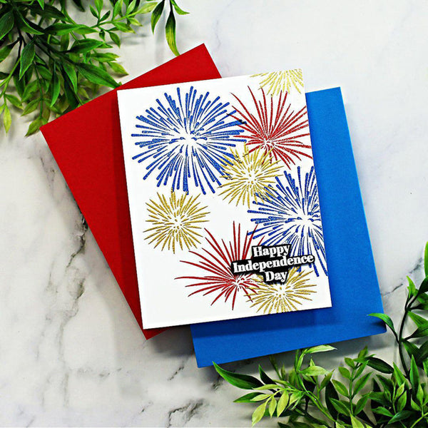 A white Independence Day greeting card with stamped red, blue, and gold fireworks, layered over red and blue envelopes on a white marble surface with green foliage.