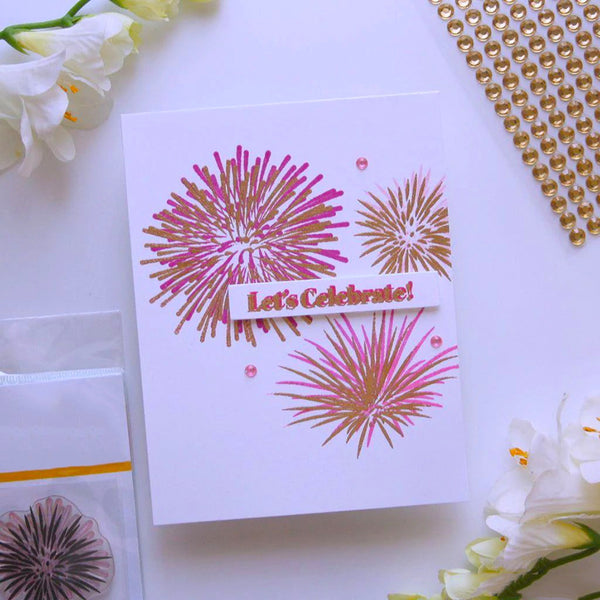 A white handmade greeting card featuring metallic pink and gold stamped fireworks and a banner that reads 'Let's Celebrate!', styled on a white surface with decorative flowers, gold beads, and a clear stamp packet.