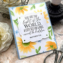 A handmade greeting card featuring a Bible verse from Matthew 5:14 in black script on a scalloped white label, surrounded by watercolor sunflowers and green leaves on a light blue card base. The card is styled on a stone surface with crafting scissors, a white rose, and a spool of thread nearby.