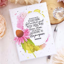 A white greeting card featuring a pink coneflower illustration and a printed Bible verse from Romans 8:28, styled on a light surface with decorative flowers, a crystal, and a pen.
