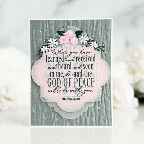 A handmade greeting card featuring a textured gray-green wood-grain background, a central scalloped pink label with a Bible verse from Philippians 4:13, and a floral embellishment at the top with pink and white flowers. The card is displayed on a white surface with blurred white flowers in the background.