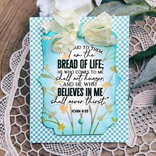 A handmade Bible verse card featuring John 6:35, styled on a lace doily with a white flower and green leaves in the background. The card has a light blue and white gingham border, a central scalloped light blue panel with yellow floral accents, and is decorated with a cream-colored ribbon bow.