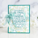 A handmade inspirational greeting card featuring a teal and white watercolor design with the Bible verse Joshua 1:9. The card is decorated with a matching teal fabric flower on the left side and is set against a soft, blurred white background.