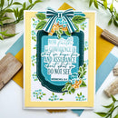 A handmade greeting card featuring a Bible verse from Hebrews 1:1, decorated with a large striped bow, green leaves, and small blue and orange flowers. The card is styled on a white surface with colorful paper and wooden props.