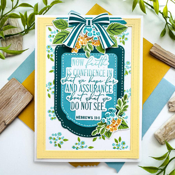 A handmade greeting card featuring a Bible verse from Hebrews 1:1, decorated with a large striped bow, green leaves, and small blue and orange flowers. The card is styled on a white surface with colorful paper and wooden props.