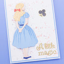 A handmade card featuring a layered die-cut illustration of Alice in a blue dress with a red bow, accompanied by a gold playing card suit symbol and the phrase 'A little magic' in gold foil, set against a light blue background.