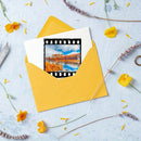 A yellow envelope on a light blue textured background, containing a white card with a printed film-strip style frame showing an autumn lakeside landscape. Yellow flowers and dried lavender sprigs are scattered around the arrangement.