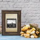 A dark brown wooden picture frame with a beige mat and a black-and-white cityscape photo insert, styled on a light blue surface against a white brick wall with yellow flowers nearby.