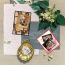 A styled flat-lay of scrapbook paper and photo frames arranged on a linen surface with green leaves and small flowers. The set includes a large white script paper, a gray cardstock sheet, a red-bordered 'Happy Day' photo frame, a black-and-tan photo frame with a vintage-style insert, and a gold scalloped shell-shaped decor piece holding small photo and postage-stamp-style cards.