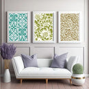 Three framed wall art prints with ornate floral and geometric patterns hang above a white sofa in a styled living room. The left print is teal and white, the center is olive green and cream, and the right is gold and cream.