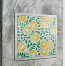 A square framed art print featuring a repeating pattern of yellow flowers and green leaves on a white background, mounted on a textured gray concrete wall.