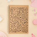 A spiral-bound notebook with a brown kraft paper cover featuring a black crackle pattern, styled in a flat-lay on a light peach background with pink stationery props.