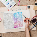A hand holds a fine paintbrush over a square art print or card featuring a geometric floral pattern. The left half of the design is white, while the right half shows a blue-to-purple-to-pink gradient.