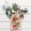 A natural burlap drawstring pouch with a red rose and green leaf print, styled with dried botanicals on a white wood background.