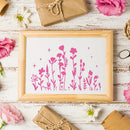 A framed art print featuring a pink silhouette of wildflowers on a white background, styled in a flat-lay with pink roses, brown kraft paper gifts, and white daisies on a distressed white wooden surface.